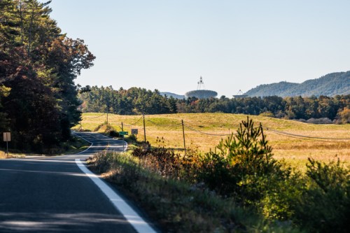 A winding road through a rural landscape with fields, trees, and distant mountains under a clear blue sky.