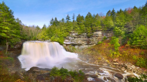 A serene waterfall cascades over rocks, surrounded by lush green trees and a clear blue sky.