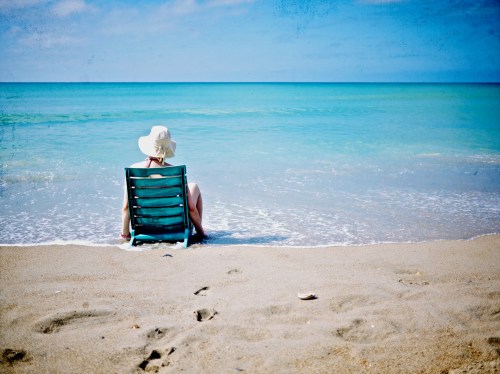 A person in a sun hat sits in a beach chair by the ocean, with gentle waves lapping at the shore.