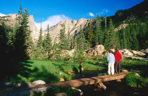 Two people stand on a wooden bridge, overlooking a lush green landscape with mountains and trees under a blue sky.