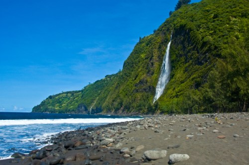 A scenic beach with smooth stones, lush green cliffs, and a waterfall cascading into the ocean under a clear blue sky.