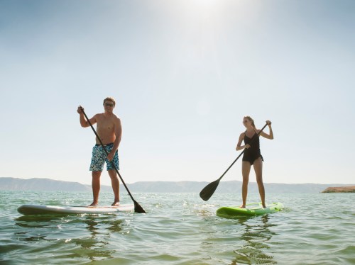 Two people paddleboarding on calm water under a bright sun, with mountains in the background.