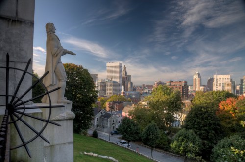 A statue overlooks a city skyline with modern buildings and greenery under a blue sky with wispy clouds.