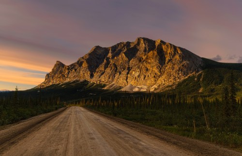 A dirt road leads towards a majestic mountain illuminated by golden sunlight under a colorful sky.