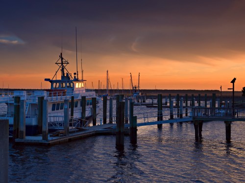 A boat docked at a marina during sunset, with calm waters and silhouettes of masts in the background.