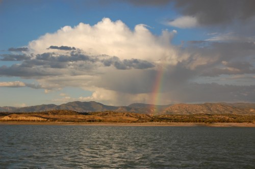 A serene landscape featuring a rainbow over mountains and a calm lake under a partly cloudy sky.