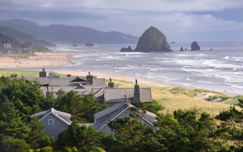 Coastal view of a beach with rocky formations, surrounded by greenery and buildings, under a cloudy sky.