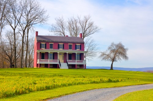 A pink historic house with a porch, surrounded by green grass and bare trees under a clear blue sky.