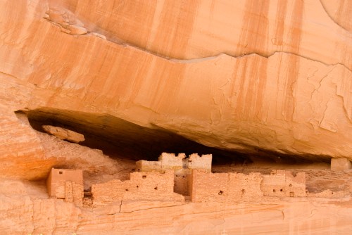 Ancient stone structures nestled under a large rock overhang in a desert landscape.