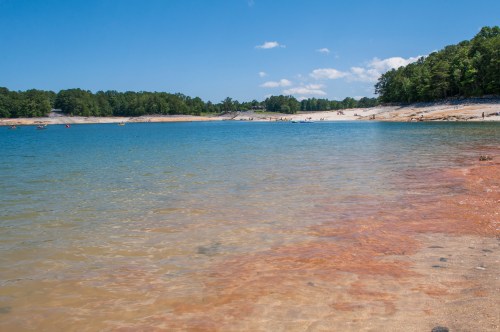A serene lake scene with clear blue water, sandy shores, and green trees under a bright blue sky.