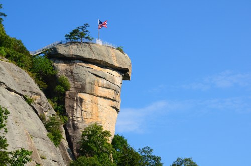 A tall rock formation topped with a flag and surrounded by greenery under a clear blue sky.
