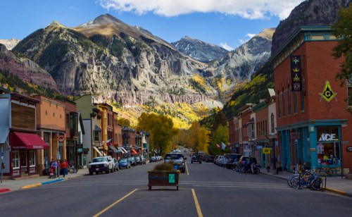 A scenic mountain town street lined with shops, trees, and parked cars, framed by towering mountains in the background.