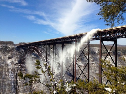 A tall bridge spans a snowy landscape, with mist or snow blowing off the edge under a clear blue sky.