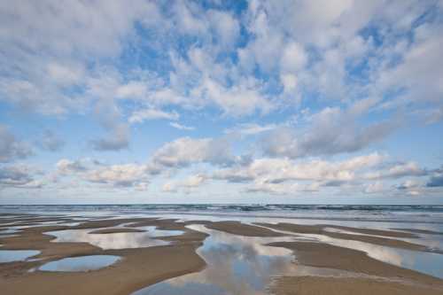 A serene beach scene with wet sand reflecting clouds and a calm ocean under a blue sky.