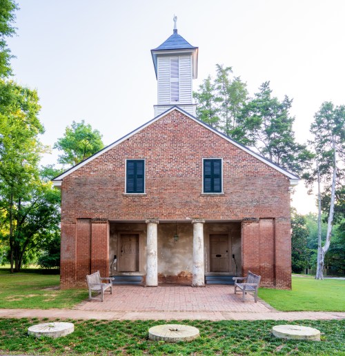 Historic brick building with a steeple, two entrances, and benches in a grassy area surrounded by trees.