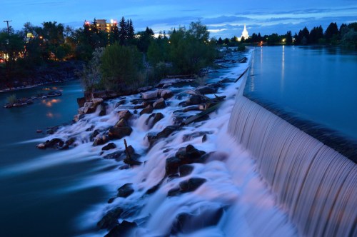 A serene river scene at dusk, featuring a waterfall cascading over rocks with trees and city lights in the background.