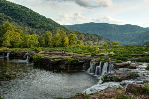 A serene landscape featuring a waterfall cascading into a calm river, surrounded by lush green hills and trees.