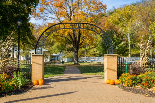 A welcoming archway at a park entrance, surrounded by colorful autumn trees and pumpkins on the path.