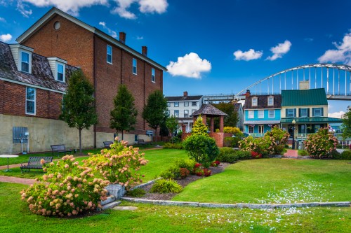 A vibrant garden with blooming flowers, benches, and historic buildings under a blue sky with clouds.