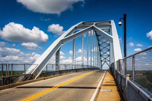 A white arch bridge spans a road under a blue sky with fluffy clouds, featuring a chain-link fence along the sides.