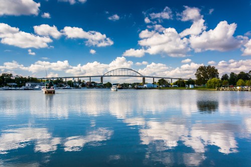 A serene river scene with boats, a bridge in the background, and fluffy clouds reflecting on the water's surface.