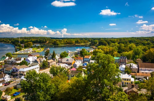A scenic view of a small town by a river, surrounded by lush greenery and dotted with boats under a blue sky.