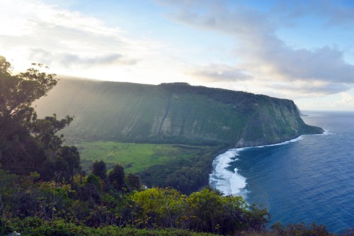 A scenic view of a lush green cliffside meeting the ocean under a cloudy sky, with sunlight illuminating the landscape.