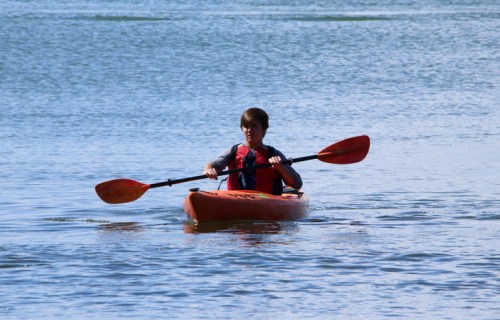 A young person paddles an orange kayak on calm water, surrounded by a serene landscape.