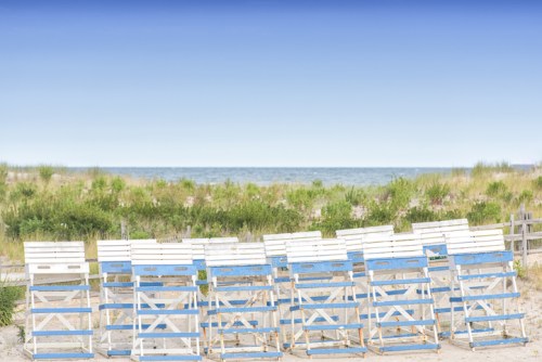 A row of white and blue beach chairs facing a calm ocean under a clear sky, surrounded by grassy dunes.