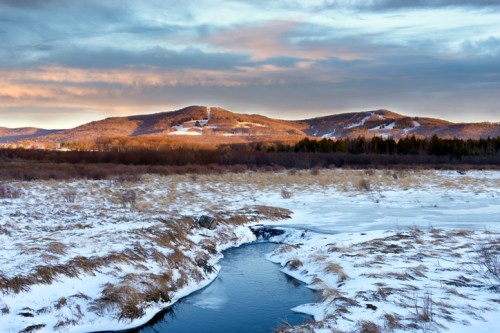 A serene winter landscape featuring a winding river, snow-covered fields, and distant mountains under a cloudy sky.