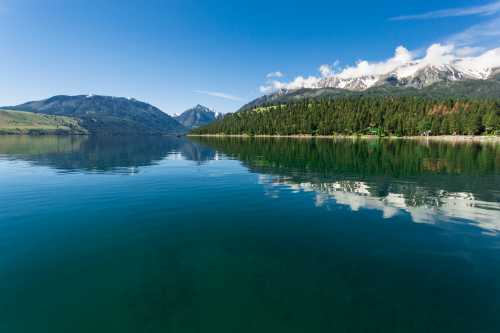 A serene lake surrounded by mountains and lush forests, reflecting the clear blue sky and clouds above.