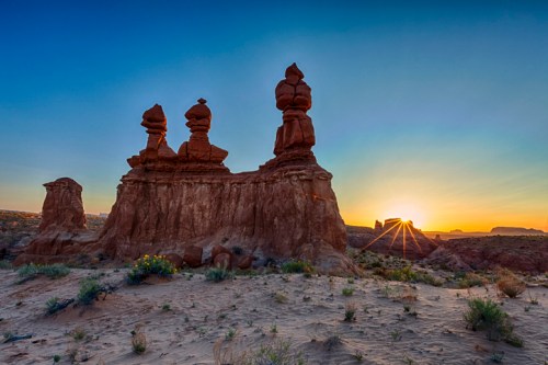Three rock formations at sunset, with warm light illuminating the landscape and desert vegetation in the foreground.