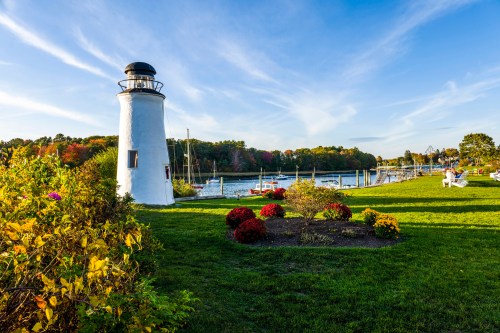 A white lighthouse stands by a calm harbor, surrounded by colorful flowers and boats under a clear blue sky.