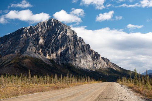 A gravel road leads towards a towering mountain under a blue sky with scattered clouds and autumn foliage in the foreground.