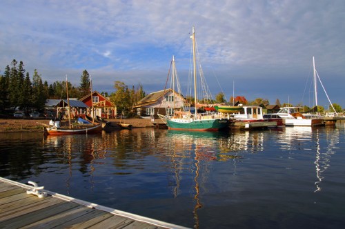 A serene harbor scene with colorful boats and reflections on calm water, surrounded by trees and a quaint building.