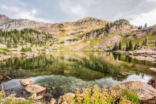 A serene mountain lake surrounded by rocky terrain and lush greenery, reflecting the cloudy sky above.