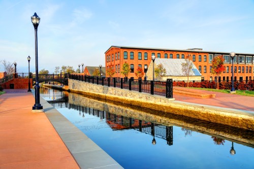 A scenic view of a canal with reflections, lined by brick buildings and lampposts under a clear blue sky.
