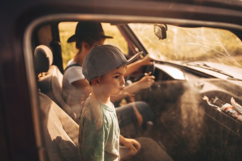 A young boy in a cap sits in a car, looking out the window, while another person drives. Sunlight filters through.
