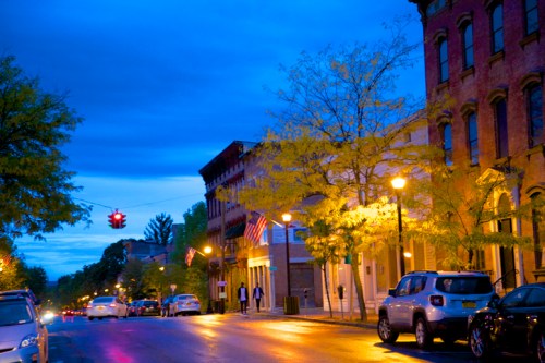 A quiet street at dusk, lined with trees and buildings, featuring streetlights and American flags.