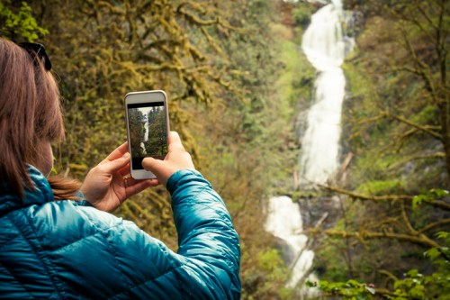 A person in a blue jacket takes a photo of a waterfall with their smartphone in a lush, green forest.
