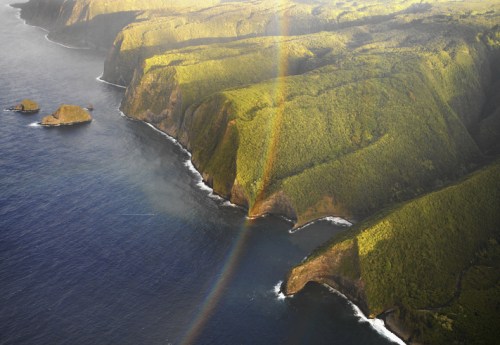 Aerial view of lush green cliffs meeting the ocean, with a vibrant rainbow arching over the landscape.