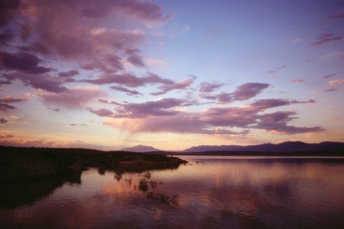 A serene lake at sunset, reflecting colorful clouds and mountains in the distance.