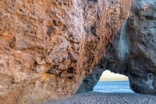 A natural rock arch on a beach, with waves gently lapping at the shore and a clear sky in the background.