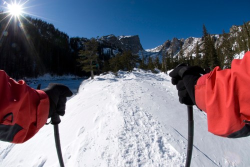 A person in a red jacket holds ski poles, looking down a snowy trail with mountains and a bright sun in the background.