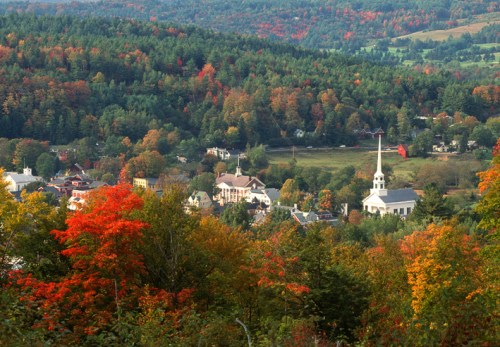 A scenic view of a small town surrounded by vibrant autumn foliage and rolling hills.