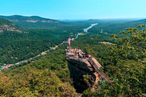 Aerial view of a rocky outcrop with people and an American flag, surrounded by lush green mountains and a winding river.