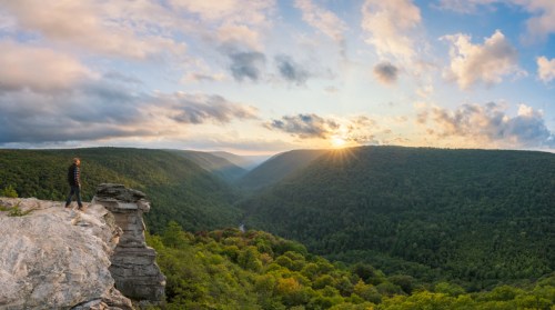 A person stands on a rocky ledge overlooking a lush green valley at sunset, with clouds scattered in the sky.