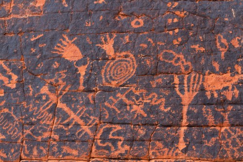 Ancient petroglyphs on a rocky surface, featuring various symbols and patterns in reddish hues.