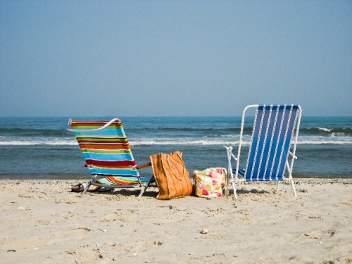 Two beach chairs on a sandy shore, with a colorful bag nearby and the ocean in the background under a clear blue sky.