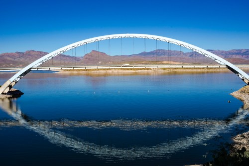 A white arch bridge spans a calm lake, reflecting in the water, with mountains in the background under a clear blue sky.
